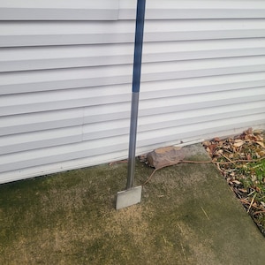May include: A long-handled, silver-colored garden tool with a black handle. The tool is leaning against a white siding wall.