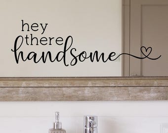 May include: A bathroom mirror with the text "hey there handsome" in black script. The mirror is framed in a light wood color and is above a white countertop with a soap dispenser, faucet, and a small mirror.