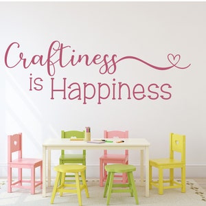 May include: A white room with a white table and colorful chairs. The wall features the phrase "Craftiness is Happiness" in pink script. The chairs are pink, green, and yellow. A rug and shelves are also visible.