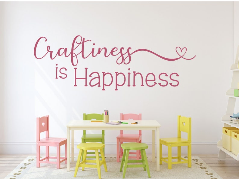 May include: A white room with a white table and colorful chairs. The wall features the phrase "Craftiness is Happiness" in pink script. The chairs are pink, green, and yellow. A rug and shelves are also visible.