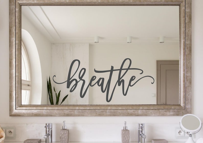 Puede incluir: Espejo rectangular con marco plateado. La palabra "breathe" est&aacute; escrita en cursiva sobre la superficie del espejo. El espejo est&aacute; sobre un lavabo de ba&ntilde;o con dos grifos y dispensadores de jab&oacute;n.