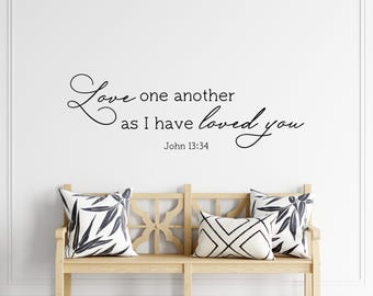 May include: A black wall decal featuring the script and serif text "Love one another as I have loved you" followed by "John 13:34" positioned above a light-colored wooden bench with three decorative pillows featuring black and white patterns.