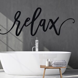 May include: A white bathtub with a grey wall behind it. The word "relax" is written in black cursive on the wall. There is a wooden stool with two bottles of soap and a stack of grey towels on it.