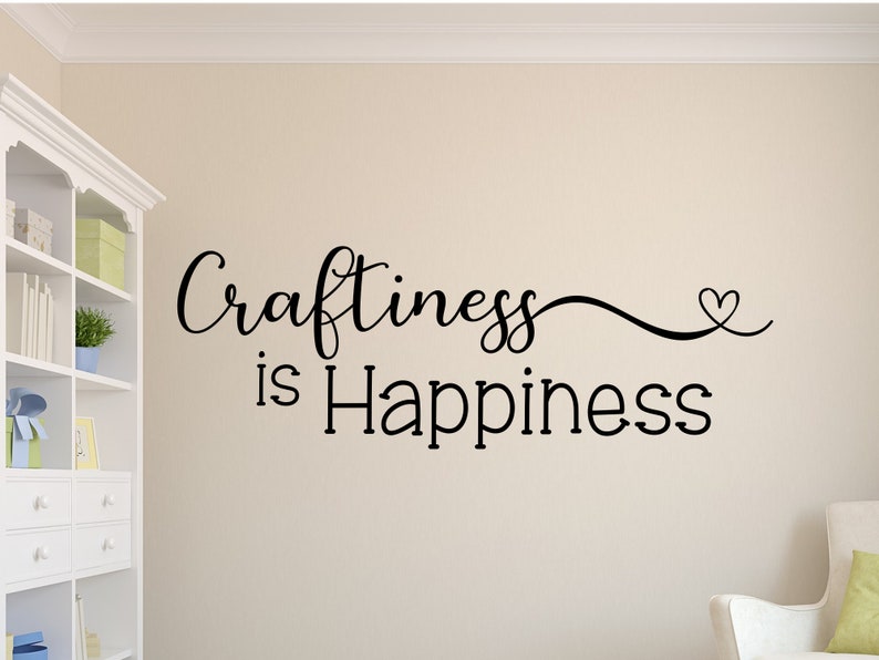 May include: Black cursive text on a beige wall reads "Craftiness is Happiness" with a heart symbol. The wall art is in a room with a white bookshelf and a white chair with a green pillow.