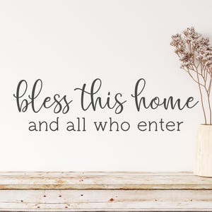 Op de afbeelding: Een witte muur met een zwarte, cursieve tekst die "bless this home and all who enter" zegt. Een vaas met gedroogde bloemen staat op een witgeschilderde houten tafel.