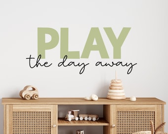 Puede incluir: Una calcomanía de pared verde claro con el texto "PLAY the day away" en letras negras. La calcomanía se muestra en una pared blanca sobre un estante de madera con un coche de juguete y un juguete de apilar de madera.