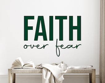 May include: A wall decal with the words "FAITH over fear" in dark green. Below, a bench holds an open book, a folded white cloth, and a string of beads. The background is a plain white wall.