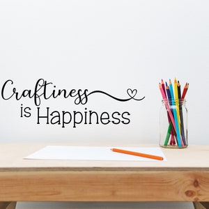 May include: A wooden desk with the phrase "Craftiness is Happiness" in black script on the wall. A clear glass jar filled with colorful pencils sits on the desk, along with a blank white paper and an orange pencil.