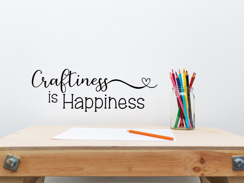 May include: A wooden desk with the phrase "Craftiness is Happiness" in black script on the wall. A clear glass jar filled with colorful pencils sits on the desk, along with a blank white paper and an orange pencil.