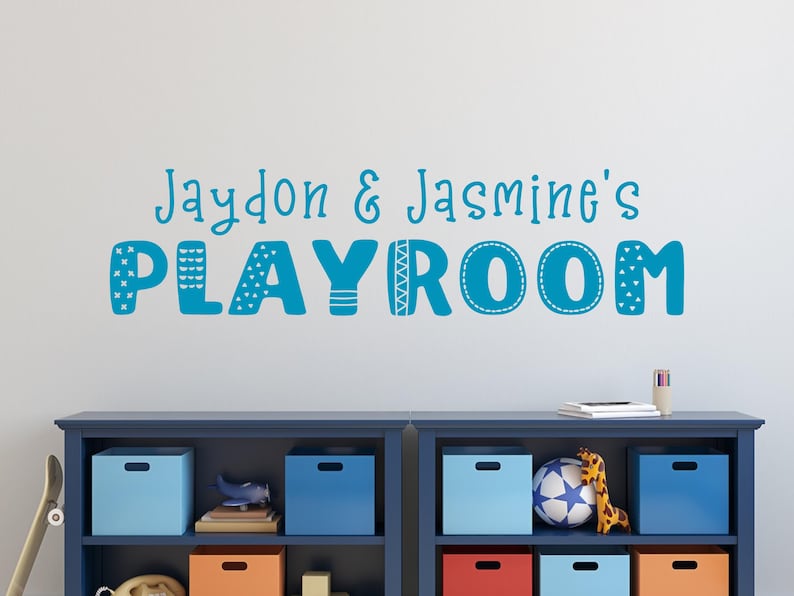 Op de afbeelding: Een blauwe en witte muursticker met de tekst "Jaydon & Jasmine's Playroom" in een speels lettertype. De sticker zit op een witte muur boven een blauwe boekenkast met kleurrijke opbergbakken.