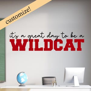 Könnte beinhalten: Ein Wandaufkleber mit dem Text "It's a great day to be a WILDCAT" in roten und schwarzen Buchstaben. Der Aufkleber befindet sich an einer weißen Wand mit einer Tafel und einem Schreibtisch im Hintergrund.
