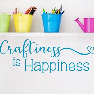 May include: A white shelf displays four colorful metal containers: a green bucket, a yellow bucket, a blue bucket, and a pink watering can. The buckets hold pencils and colored pencils. The phrase "Craftiness is Happiness" is written in teal.