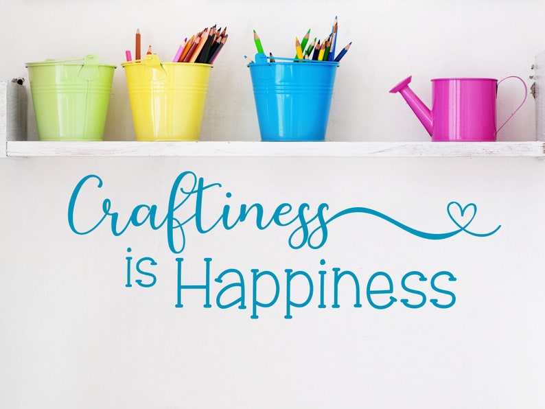 May include: A white shelf displays four colorful metal containers: a green bucket, a yellow bucket, a blue bucket, and a pink watering can. The buckets hold pencils and colored pencils. The phrase "Craftiness is Happiness" is written in teal.
