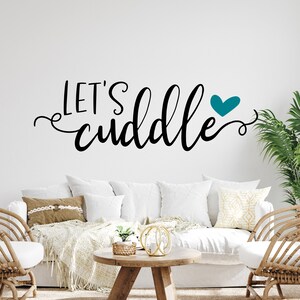 Könnte beinhalten: Ein weißes Sofa mit Kissen und einer Decke in einem Wohnzimmer. Die Wand hinter dem Sofa hat einen schwarzen und weißen Vinyl-Aufkleber mit der Aufschrift "Let's cuddle" und einem türkisfarbenen Herz.