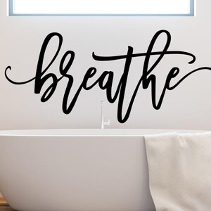 Puede incluir: Adhesivo de pared negro con la palabra "breathe" en escritura cursiva sobre una ba&ntilde;era blanca. Una toalla blanca cuelga del borde de la ba&ntilde;era. El ba&ntilde;o tiene un dise&ntilde;o minimalista.