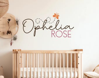 May include: A nursery wall decal featuring the name "Ophelia Rose" in a stylish script font, with a butterfly accent. The design is in brown and mauve colors, above a wooden crib.