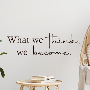 May include: A white wall with a brown inspirational quote that reads "What we think, we become." The quote is written in a cursive font. There is a wicker chair with a white throw blanket and a stack of books on a small wooden table in the foreground.