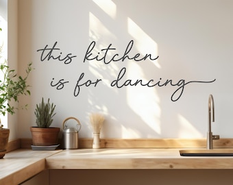 May include: A kitchen scene with the text "this kitchen is for dancing" in a cursive font on a white wall. A wooden countertop features a sink, plants, and decorative items. Natural light creates shadows.