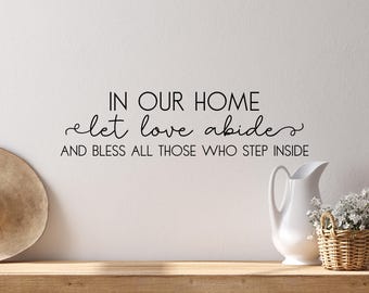 May include: A wall decal with the text "IN OUR HOME let love abide AND BLESS ALL THOSE WHO STEP INSIDE" in black script. A white pitcher, a small wicker basket with flowers, and a straw hat are on a wooden shelf.