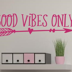 May include: Pink vinyl wall decal with the text "GOOD VIBES ONLY" and a pink arrow with a heart in the middle. The decal is on a white wall above a set of three drawers.