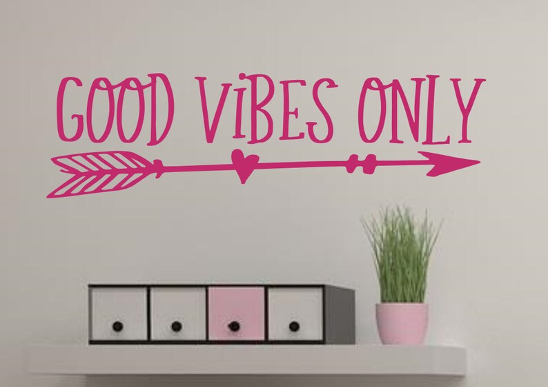 May include: Pink vinyl wall decal with the text "GOOD VIBES ONLY" and a pink arrow with a heart in the middle. The decal is on a white wall above a set of three drawers.