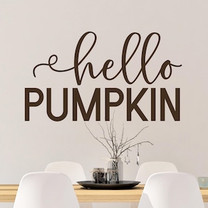 May include: A brown wall decal with the text "hello pumpkin" in a cursive font. The decal is displayed on a white wall above a wooden table with white chairs.