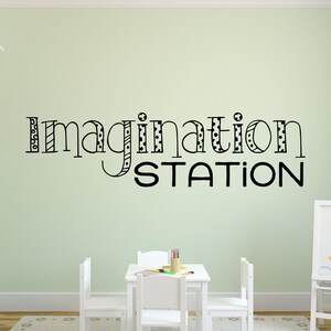 May include: A black and white wall decal with the words "Imagination Station" in a playful font. The decal is on a light green wall in a room with white furniture and two white chairs.