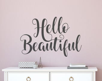 May include: A wall sticker featuring the phrase "Hello Beautiful" in dark grey cursive lettering. The text is set against a pale pink wall. A white chest of drawers with gold handles and decorative items is below.