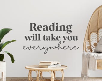 May include: A white wall with a black vinyl decal that reads "Reading will take you everywhere". A wicker chair with a white throw blanket is in the background. A small wooden table with a stack of books sits in the foreground.