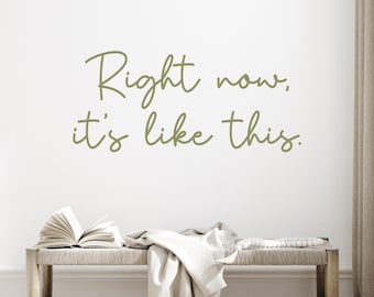 May include: A wall with the text "Right now, it's like this" in a cursive, olive green font. Below the text is a wooden bench with an open book, a white cloth, and a beaded garland.