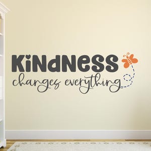 May include: A wall decal with the text "Kindness changes everything" in black lettering with a small heart above the "d" in kindness. A small orange butterfly with a blue dotted line trailing behind it is to the right of the text.