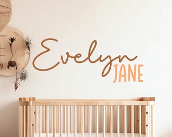 May include: A white wall with a brown and orange cursive name sign that reads "Evelyn Jane" above a wooden crib.