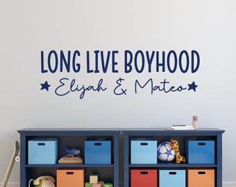 May include: A navy blue wall decal with the text "LONG LIVE BOYHOOD" and the names "Elijah & Mateo" in a script font. The decal is above a blue storage unit with colorful storage boxes and toys.