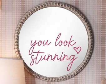 May include: A round mirror with a distressed, beaded frame. The mirror's surface displays the words "you look stunning" in a cursive, magenta font, accompanied by a heart symbol. The mirror is mounted on a patterned wall.