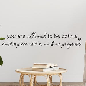 May include: A white wall with a black cursive quote that reads "You are allowed to be both a masterpiece and a work in progress" with a small heart symbol at the end. The quote is centered on the wall. There is a small potted plant on the left side of the image and a wicker chair on the right side of the image.