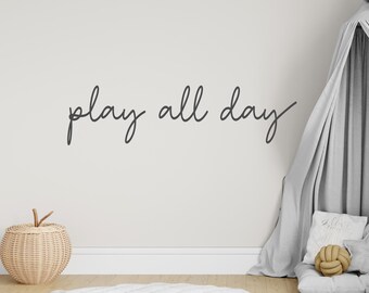 Puede incluir: Una escena de guardería en tonos neutros con la frase "play all day" en escritura cursiva en una pared blanca. Una cesta de mimbre y un dosel gris completan el ambiente acogedor.
