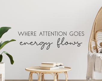 May include: A white wall with the text "Where attention goes energy flows" written in black. A wicker chair with a white throw blanket is on the right side of the image. A small wooden table with a stack of books is in the center of the image. A green plant is on the left side of the image.