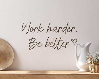 May include: A motivational wall art piece with the text "Work harder. Be better" in a handwritten style. A white pitcher with flowers and a woven basket are on a wooden shelf, with a straw hat on the left.