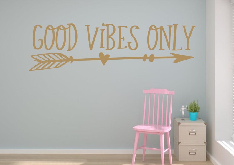 May include: A gold wall decal with the text "GOOD VIBES ONLY" and an arrow with hearts. The decal is on a light blue wall with a pink chair and a small table with a plant on it.