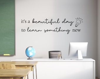 May include: A black wall decal featuring the text "it's a beautiful day to learn something new" in a mix of serif and script fonts, accompanied by a simple line-art illustration of a lightbulb, applied to a plain white wall surface.