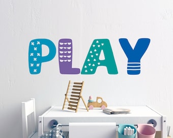 Pode incluir: Um adesivo de parede colorido que soletra "PLAY" com padrões diferentes em cada letra. As letras são azuis, roxas, azul-turquesa e azuis. O adesivo está em uma parede branca acima de uma mesa branca com um ábaco de madeira e um caminhão de brinquedo.