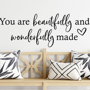 Op de afbeelding: Een witte houten bank met drie kussens. De kussens hebben zwart-witte geometrische patronen. Een zwarte vinyl wand sticker met de tekst "You are beautifully and wonderfully made" en een klein hartje is op de muur achter de bank.