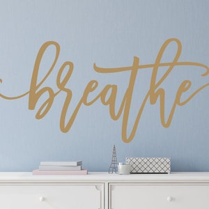 Puede incluir: Una calcoman&iacute;a de pared dorada con la palabra "breathe" en una elegante escritura se muestra en una pared azul claro. La palabra es el punto focal, con una c&oacute;moda blanca y objetos decorativos debajo.