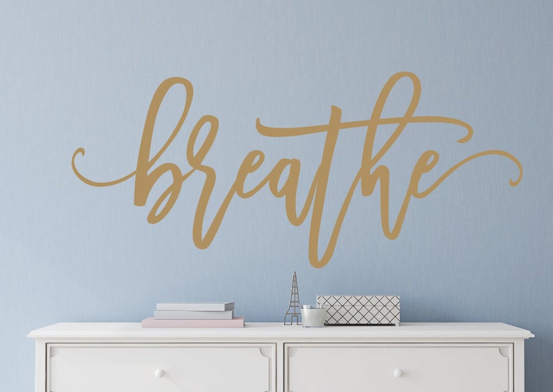 Puede incluir: Una calcoman&iacute;a de pared dorada con la palabra "breathe" en una elegante escritura se muestra en una pared azul claro. La palabra es el punto focal, con una c&oacute;moda blanca y objetos decorativos debajo.