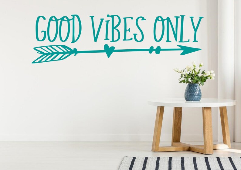 May include: Teal wall decal with an arrow and the text "GOOD VIBES ONLY". The decal is on a white wall with a white table and a vase of white flowers.