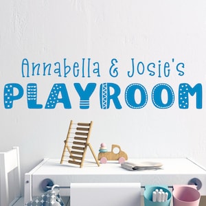 Op de afbeelding: Een blauw en wit speelkamerbord met de tekst "Annabella & Josie's Playroom" op een witte muur. Het bord heeft een speels lettertype met decoratieve elementen.