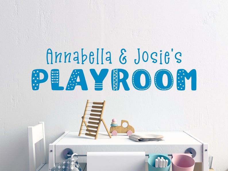 Op de afbeelding: Een blauw en wit speelkamerbord met de tekst "Annabella & Josie's Playroom" op een witte muur. Het bord heeft een speels lettertype met decoratieve elementen.