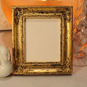 May include: A gold-colored picture frame with ornate detailing. The frame has a distressed finish and is made of wood.