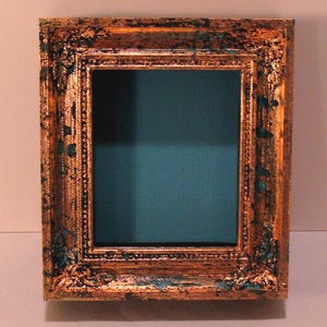 May include: Ornate, rectangular picture frame with a gold finish and teal accents. The frame has detailed carvings and a dark teal interior, creating a vintage aesthetic. Ideal for displaying photos or artwork.