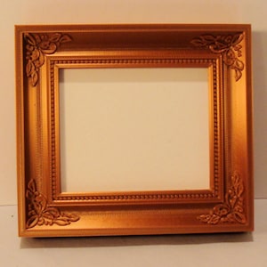 May include: A rectangular gold-colored picture frame with ornate corner detailing. The frame has a beaded inner border and a smooth outer edge. The corners feature floral-like embellishments, adding a decorative touch to the frame's design.